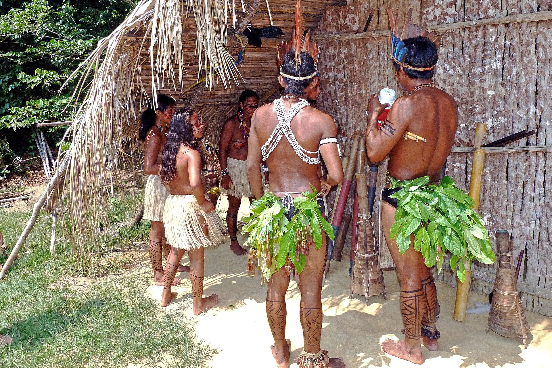 Das Bild zeigt eine Gruppe von Amazonas Indianern vor einer traditionellen Hütte aus Holz und Bambus. Die Frauen tragen bunte Baströcke und die Männer grüne Blätter als "Hose".