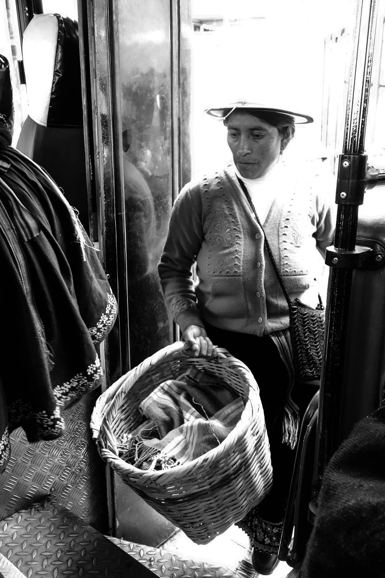 Eine schwarz-weiß Fotografie einer indianischen Frau aus Südamerika, die einen Hut und ein Kleid trägt und eine Strickweste darüber an hat. Sie geht in ein Geschäft mit einem Weidenkorb, der mit Wäsche gefüllt ist.