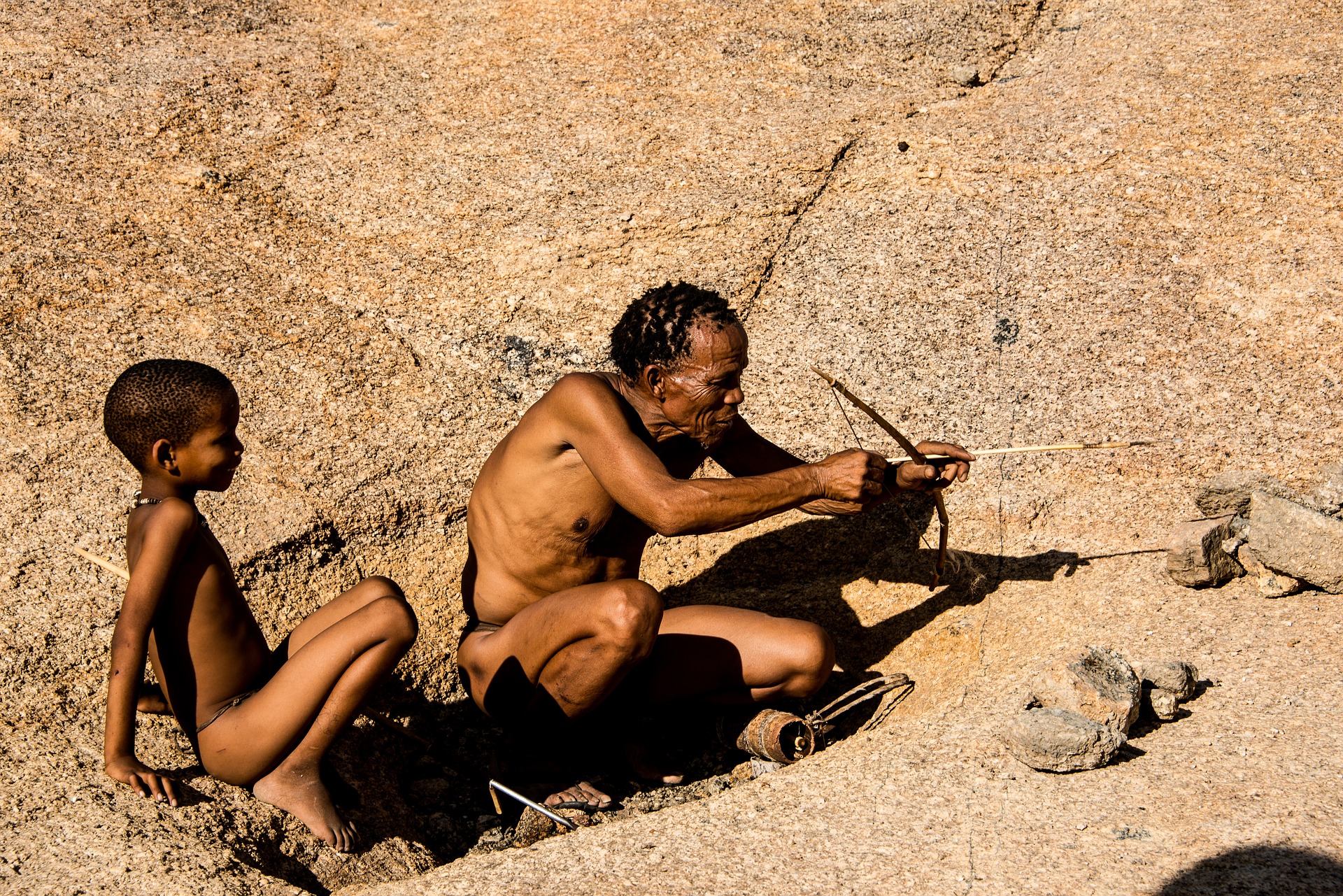 Ein älterer Indianermann im Lendenschurz sitzt auf einem Felsen und hält einen Bogen und Pfeile in den Händen. Neben ihm sitzt ein kleiner Junge und schaut ihn gespannt an. Die Landschaft im Hintergrund ist kahl und öde.