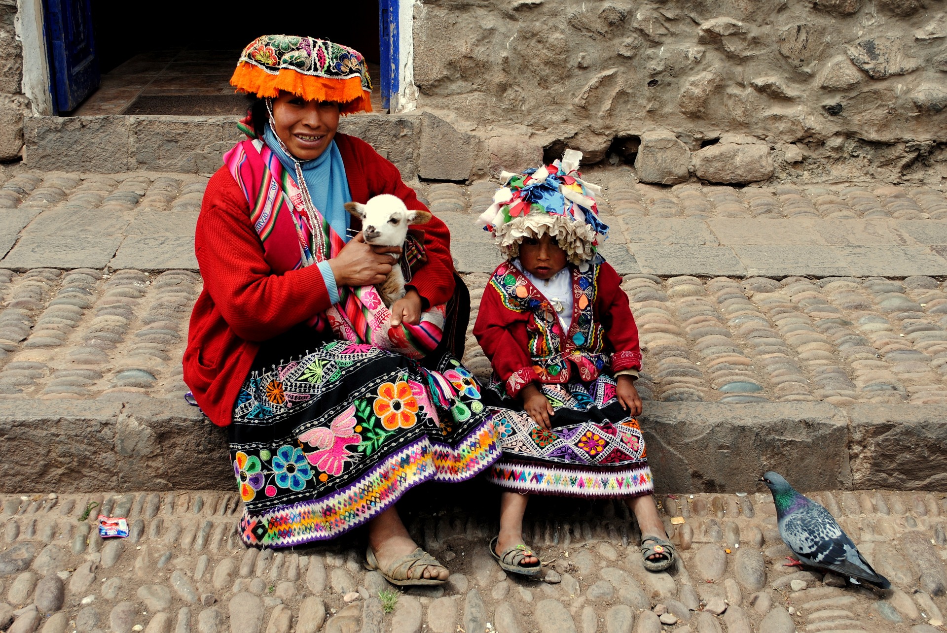 Mutter und Kind der Peru-Indigenen in traditioneller bunter Kleidung, sitzen auf einem Gehsteig. Die Mutter hält eine Ziege in ihren Armen, während das Kind neben ihr sitzt. Die Mutter lächelt in die Kamera, während das Kind erstaunt und verwundert in die Kamera schaut.