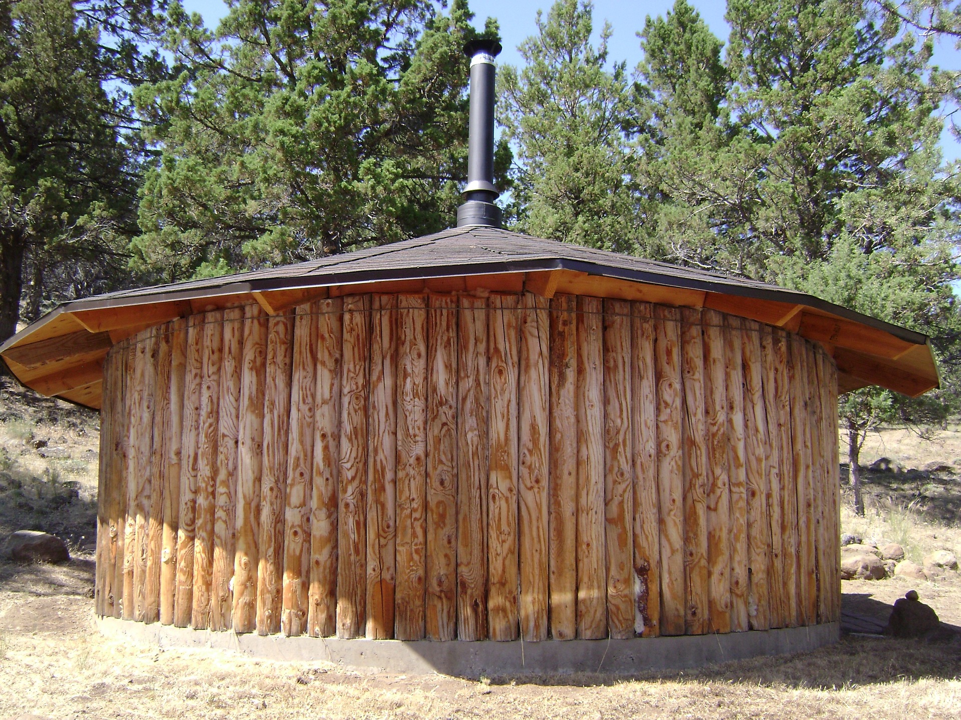 Nahaufnahme einer runden Schwitzhütte aus Holz, die von den indianischen Stämmen zur Reinigung verwendet wird. Das Holz ist dunkelbraun und das Dach hat eine Kuppelform. Die Hütte steht auf Sand und im Hintergrund sind Nadelbäume zu sehen.
