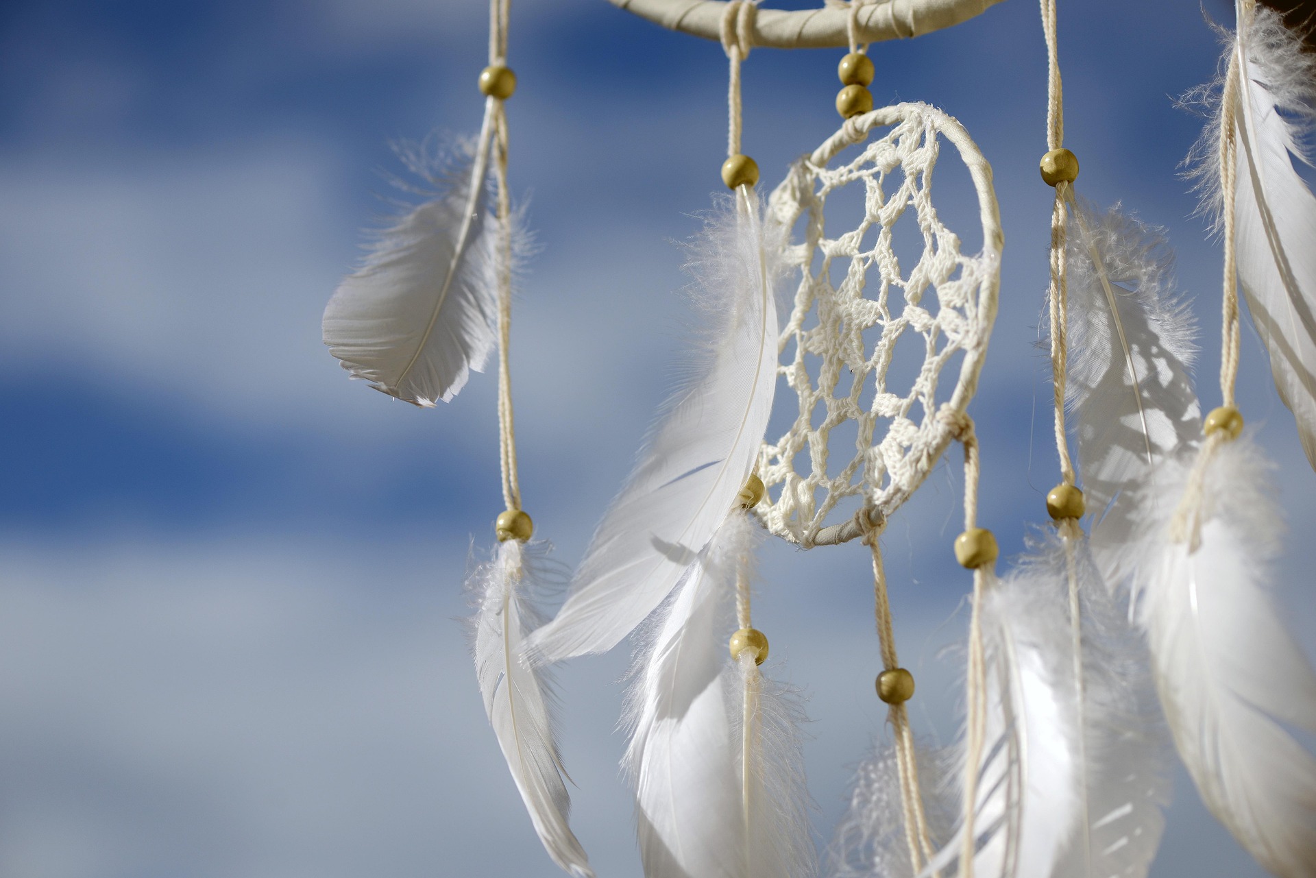 Weißer Traumfänger mit Federn und Perlen, auf blauem Himmel mit Wolken im Hintergrund. Der Ausschnitt zeigt das Netz des Traumfängers.